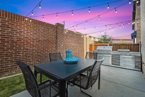 Eine Terrasse mit Backsteinmauer, Tisch mit Stühlen, Lichterkette und eingebautem Grill.