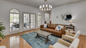 Ein geräumiges Wohnzimmer mit einem braunen Ledersofa, einem Couchtisch und Korbstühlen. Große Fenster mit Rundbögen und ein Kronleuchter sind vorhanden.