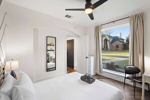 Quarto de hotel com uma cama grande, ventoinha no teto, escrivaninha com cadeira, janela com cortinas e vista para um edifício.