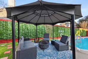 Uma área de estar coberta ao ar livre com lareira, espreguiçadeiras e uma piscina ao fundo.