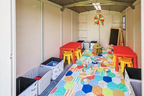 Ein Kinderraum mit Tischen, Stühlen und einer bunten Spielmatte.