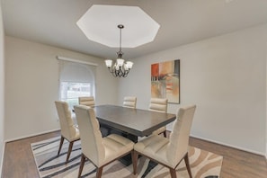 A dining area with a dark wooden table, six beige chairs, a chandelier, a window with blinds, and a painting on the wall. - Spacious and Modern Home With Private Pool (Plano)