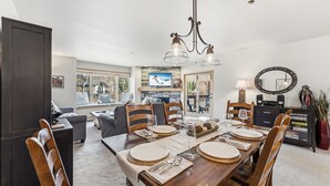 A dining area with a wooden table set for six, a television mounted on the wall, and a chandelier. - Breck Getaway | Trails, Balcony & Shared Hot Tub (Breckenridge)