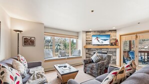A living room with a sofa, armchair, and a coffee table. There is a TV mounted on the wall and a window with a view of the outdoors. - Breck Getaway | Trails, Balcony & Shared Hot Tub (Breckenridge)
