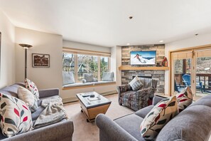 A living room with a sofa, armchair, and a coffee table. There is a TV mounted on the wall and a window with a view of the outdoors. - Breck Getaway | Trails, Balcony & Shared Hot Tub (Breckenridge)