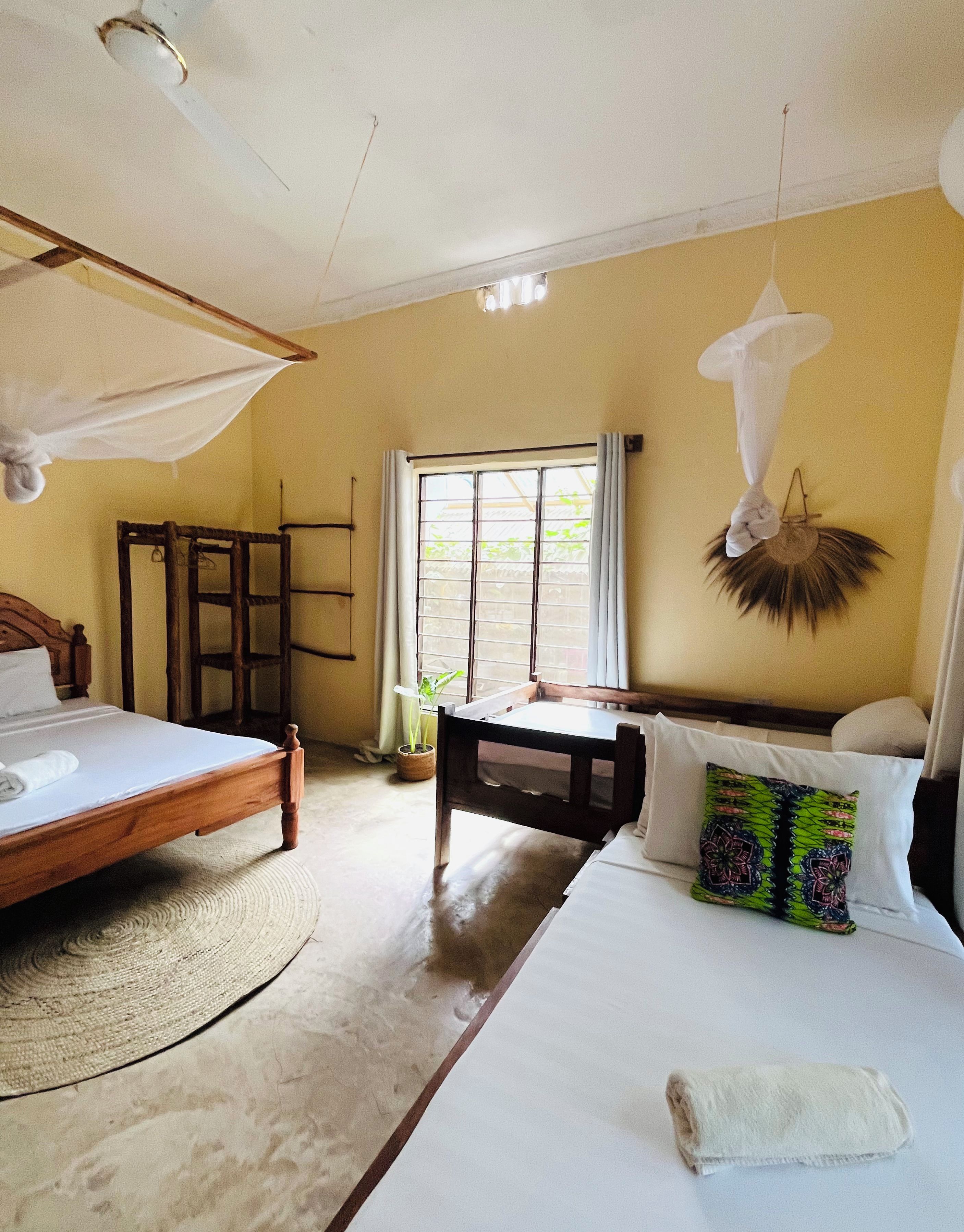A room with a bed, a bedside table, a window with curtains, a hanging mosquito net, and a woven mat on the floor.