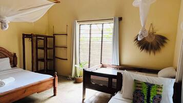 A room with a bed, a bedside table, a window with curtains, a hanging mosquito net, and a woven mat on the floor.