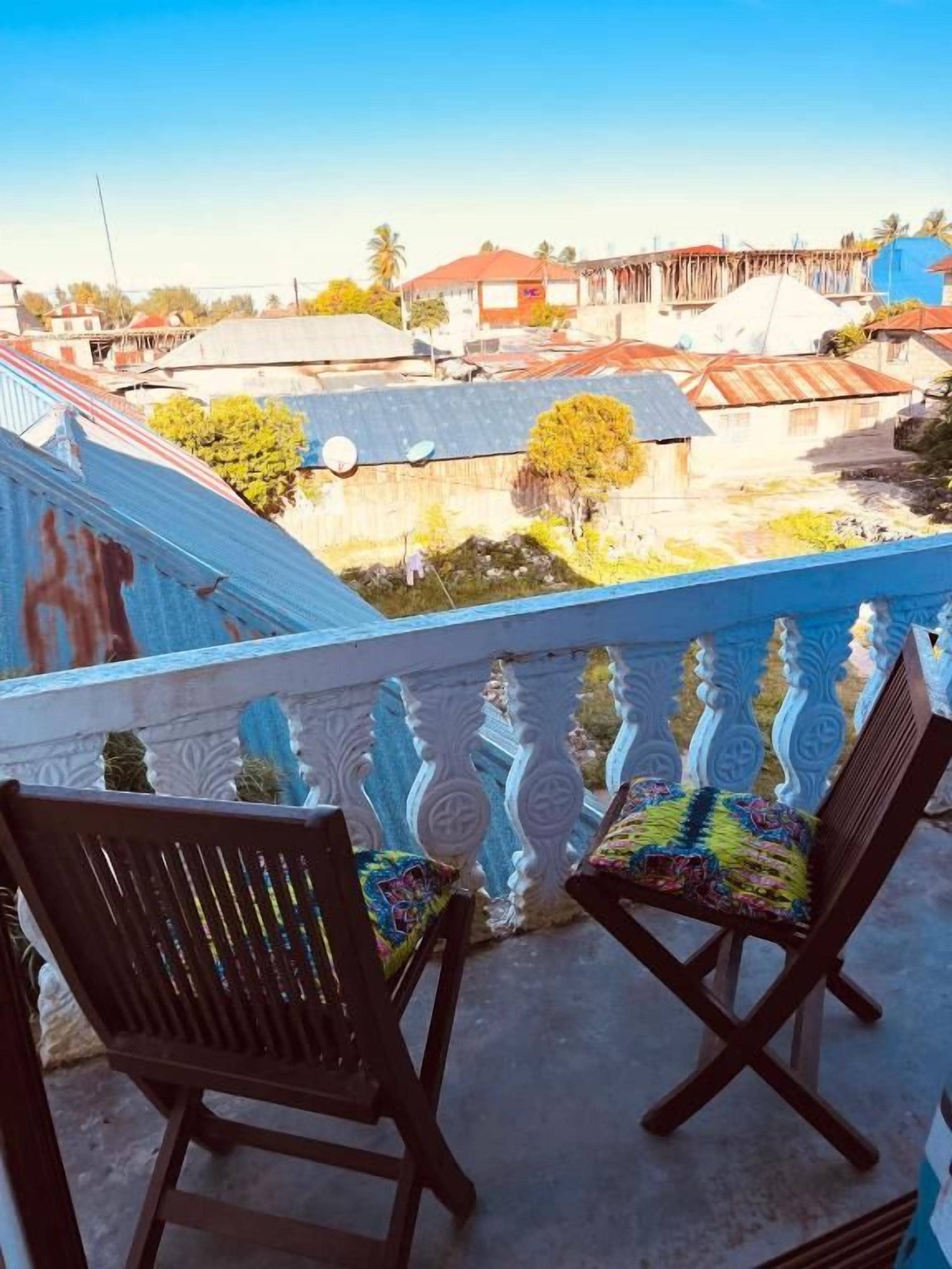 Un balcone con due sedie di legno, una ringhiera con elementi decorativi e vista su una zona residenziale con case e alberi.