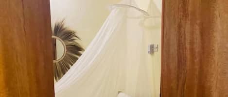 Una habitación con una cama bajo un dosel blanco, un espejo redondo en la pared y una puerta de madera.
