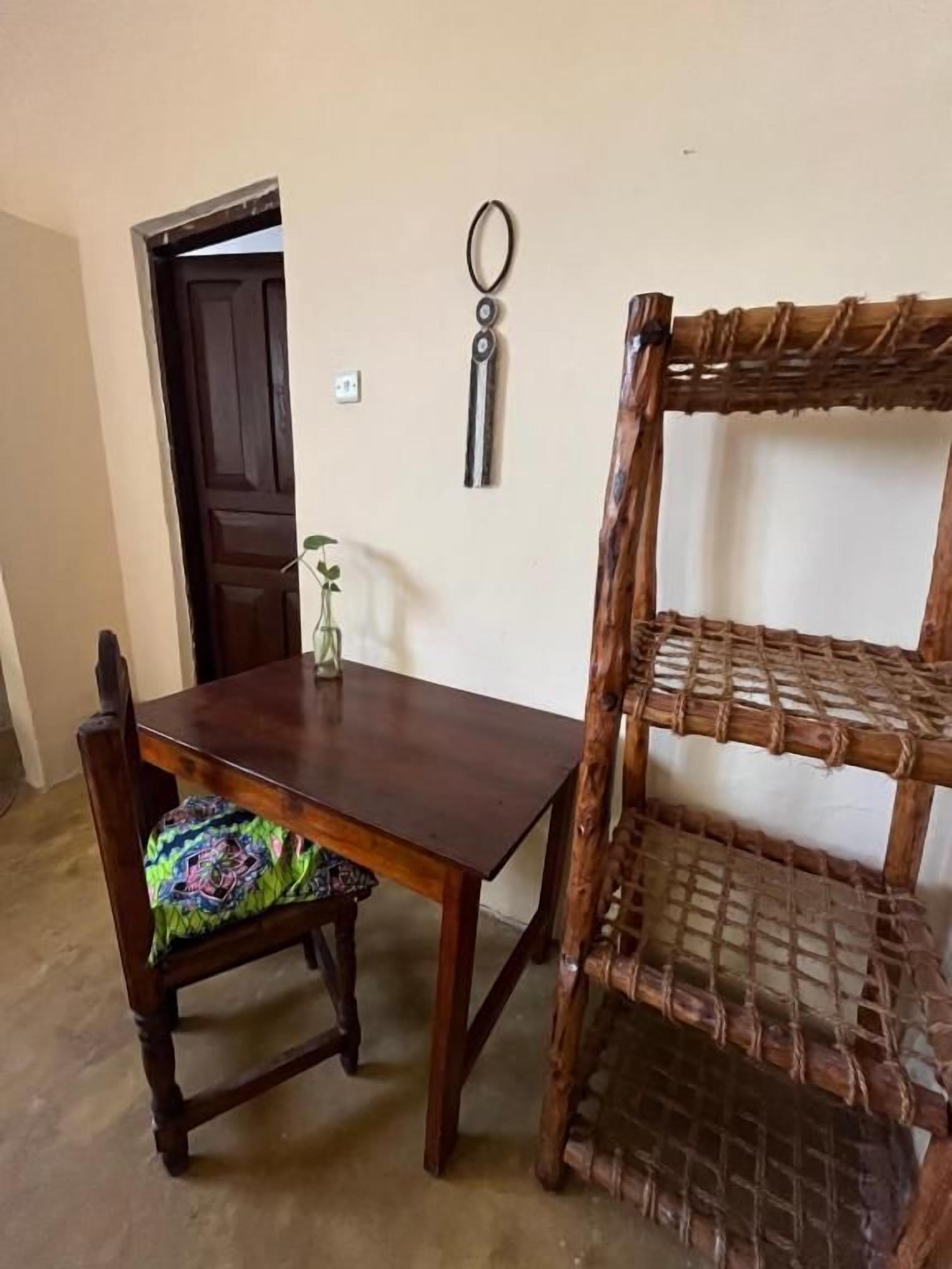 Une table à manger en bois, accompagnée d’une chaise, d’une étagère en osier et d’une porte visible au fond.