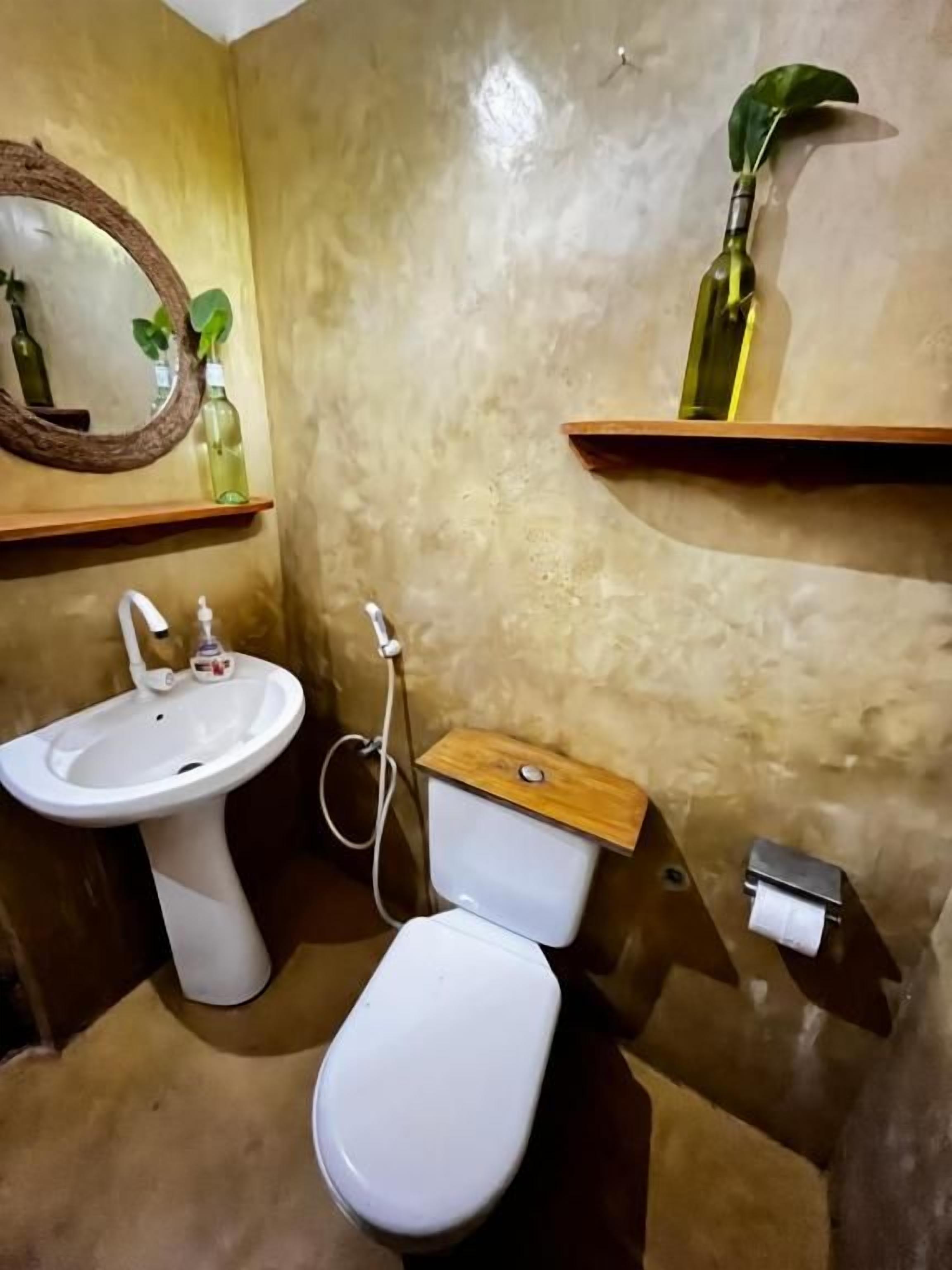 Et badeværelse med hvid vask, toilet og en træhylde med en flaske.