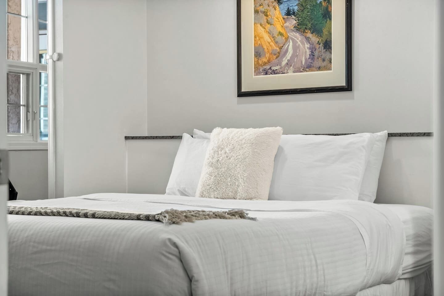 A bedroom with a bed, white bedding, a painting on the wall, and a window with blinds.
