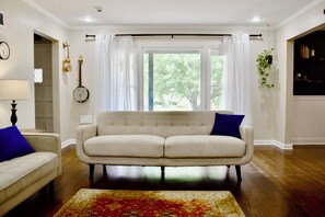 A living room with a sofa, a rug, a lamp, a guitar, a banjo, and a large window with curtains.