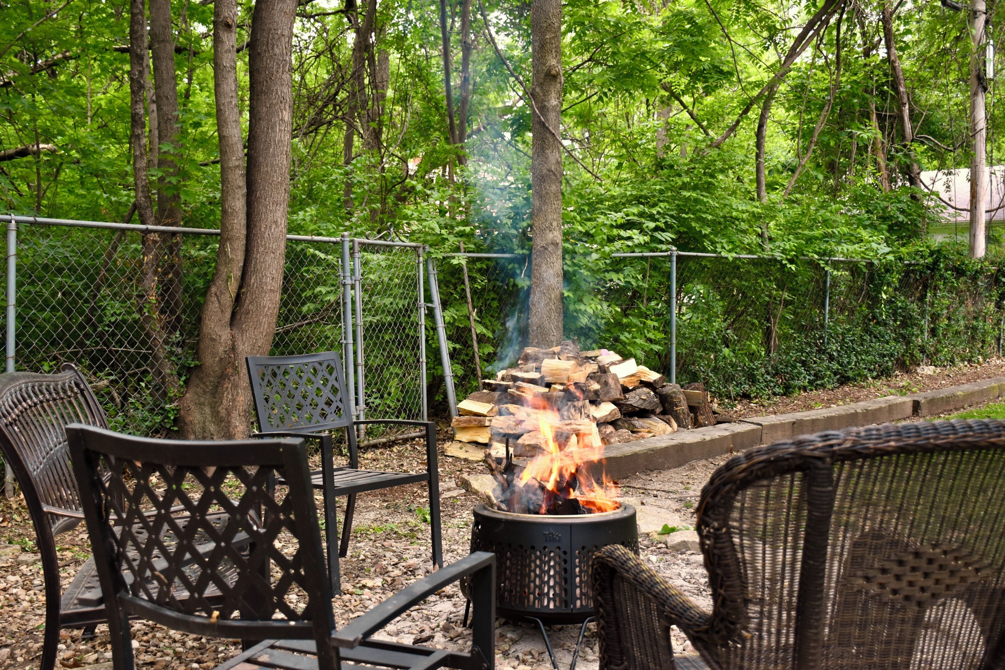 A fire pit with logs, surrounded by wicker chairs, in a wooded outdoor area.