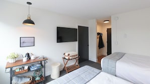 A hotel room with a bed, a desk with a kettle, a fan, a TV, and a door leading to another room.