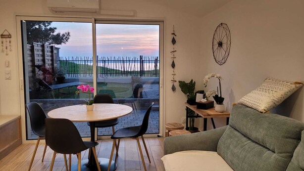 Een kamer met een schuifdeur naar een balkon, een ronde tafel met stoelen, een bank, en een klein houten tafeltje met planten en een zakdoosje.