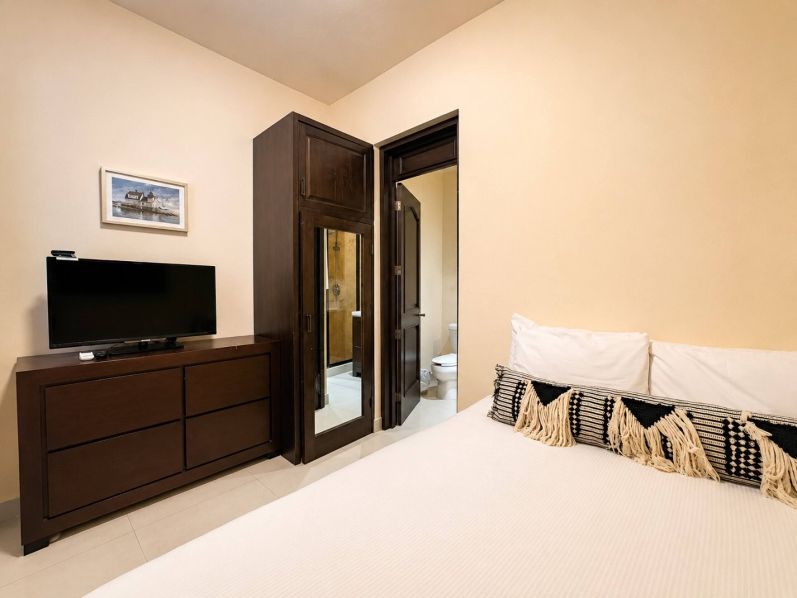 A bedroom with a bed, a TV on a dresser, a door leading to a bathroom, and a framed picture on the wall.