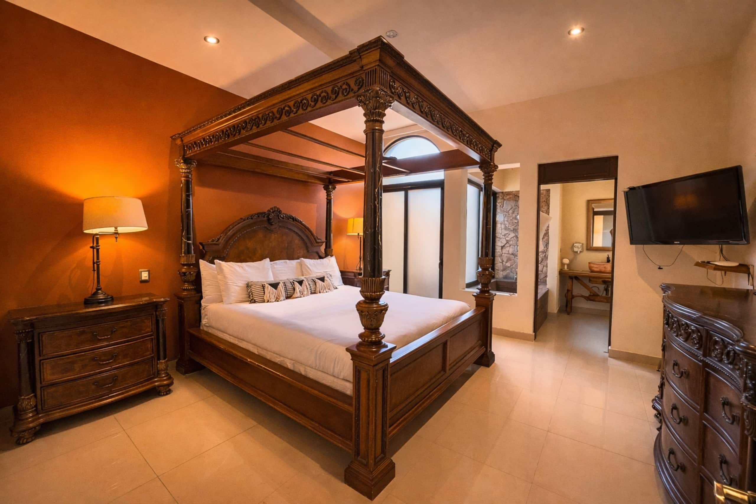 A four-poster bed with a wooden frame and white bedding, a nightstand with a lamp, a dresser with drawers, and a television on the wall.