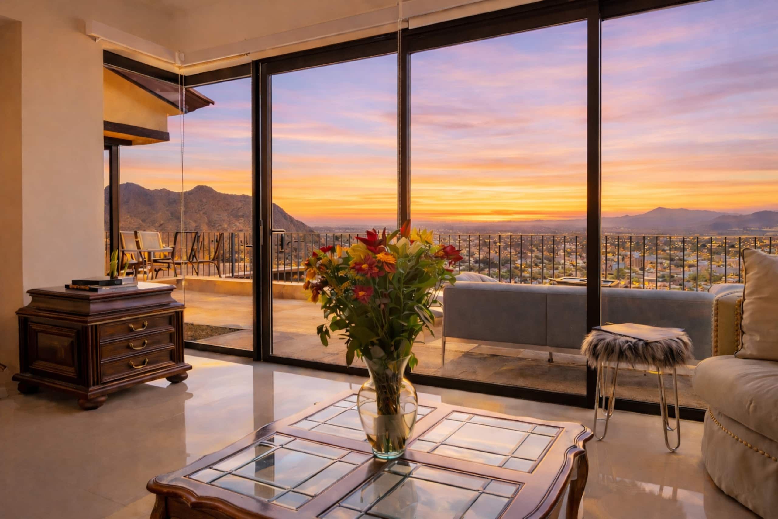 A modern living room with a large glass table, a sofa, and a view of a cityscape at sunset.