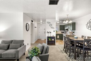 A modern living room with a grey sofa, a dining area with a wooden table and chairs, and a kitchen in the background. - Casa de Knight 2.0 (Las Cruces)