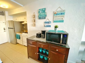 A kitchen with a refrigerator, stove, microwave, and cabinets. There are wall decorations with phrases like 'Sea & Sunshine' and 'Shoes Optional.'