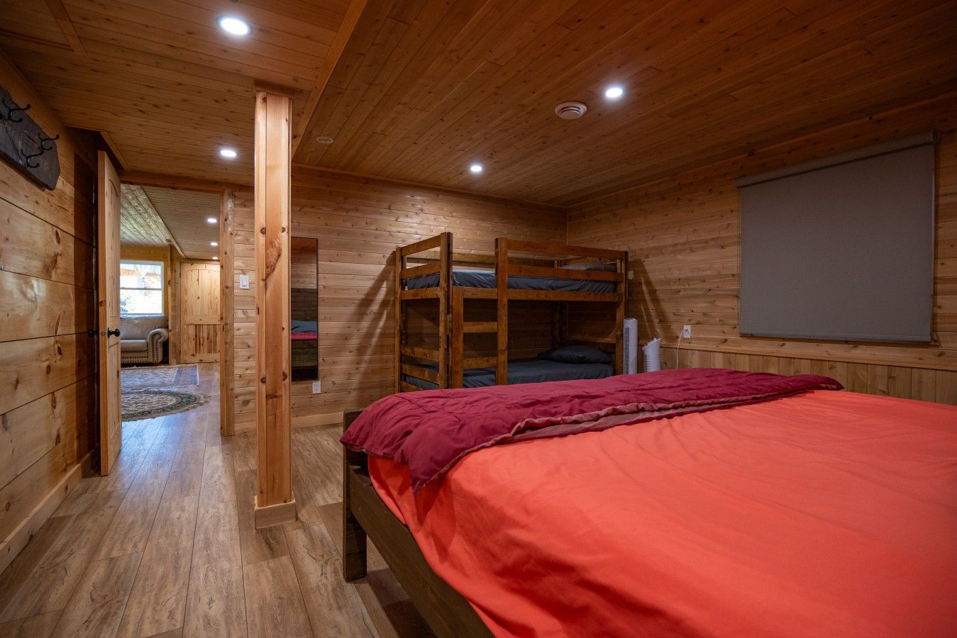 A wooden cabin room with a bunk bed, a single bed with a red blanket, and a window with a view.