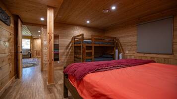 A wooden cabin room with a bunk bed, a single bed with a red blanket, and a window with a view.