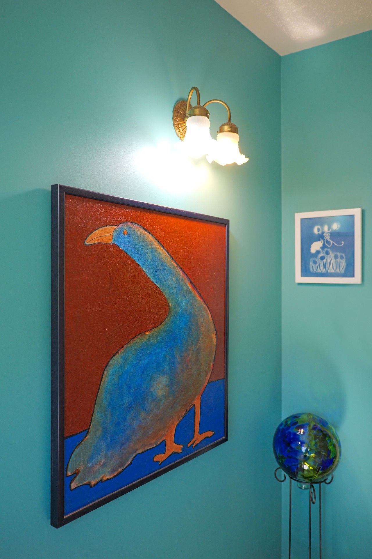 Una pared color turquesa con un cuadro enmarcado de un pájaro, un accesorio de luz fijado a la pared y un pequeño globo terráqueo sobre un soporte.