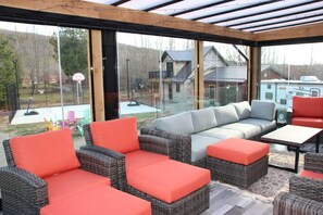 Een terras buiten met rieten meubels en kussens, een glazen overkapping en uitzicht op huizen en een basketbalring.