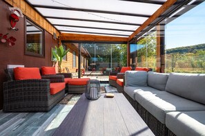A modern outdoor seating area with wicker furniture and cushions, a glass enclosure, and a view of the landscape.