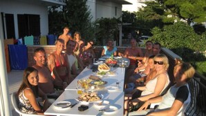 A group of people seated around a table with food and drinks, outdoors with a building and trees in the background. - Seaview Family Apartment With Grand Terrace (Omis)