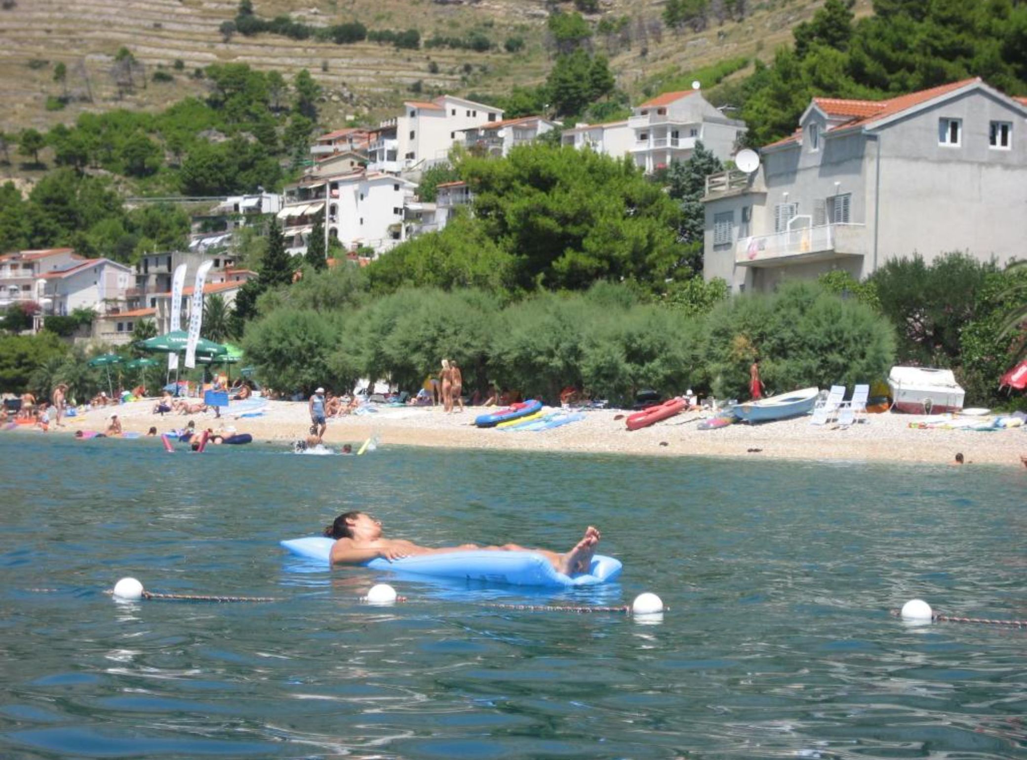 Une personne flottant sur un matelas gonflable bleu dans une eau calme, une plage avec des gens et des bateaux, et des bâtiments en arrière-plan.