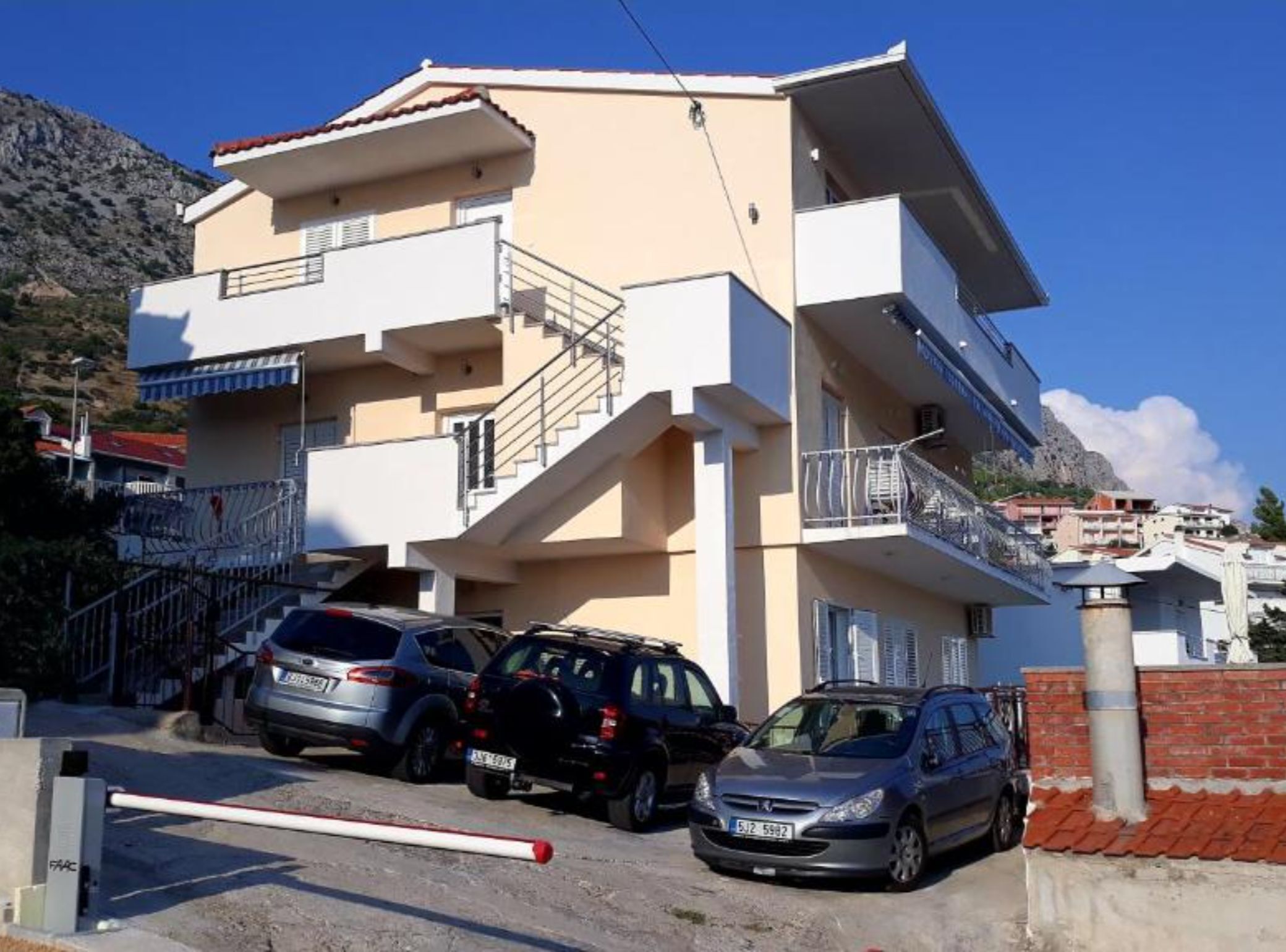 A multi-story residential building with balconies, parked cars, and a mountainous backdrop.