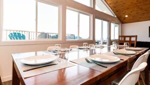 A dining area with a long wooden table set for multiple people, large windows, and a view of the outdoors.