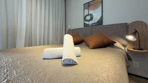 Ein ordentlich bezogenes Bett mit aufgerollten Handtüchern, einem Kopfteil, einem Nachttisch mit Lampe und einem an der Wand befestigten abstrakten Kunstwerk.