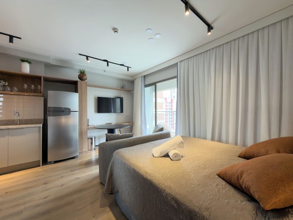 Ein modernes Hotelzimmer mit Bett, Schreibtisch, Stuhl, Küchenzeile und einem großen Fenster.