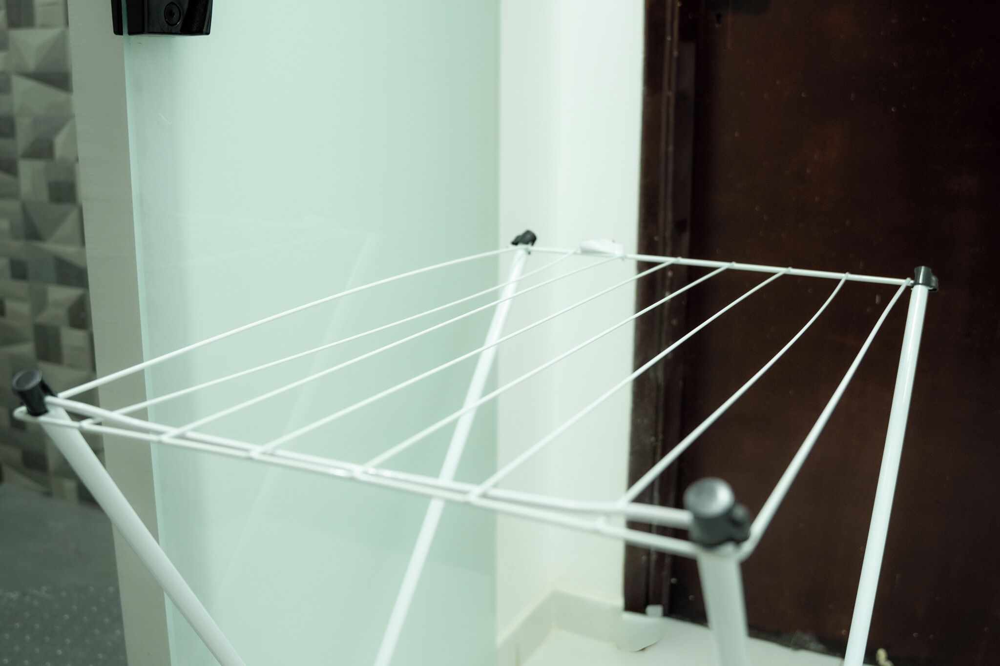 A metal drying rack with a white frame, mounted on a wall with a dark door and a tiled wall in the background.