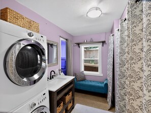 A laundry area with a front-loading washer and dryer, a sink, a window with a view of the outside, and a blue cushioned bench.