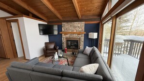 Un salón con chimenea de piedra, un sillón de cuero, un sofá modular gris y un televisor montado en la pared.