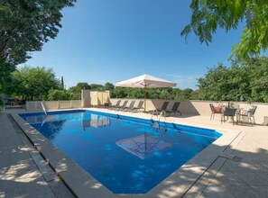 A rectangular swimming pool with a clear blue water surface, surrounded by a tiled deck, lounge chairs, and a table with chairs. - Villas Casa Bianca & Maria Residence (Marcana)