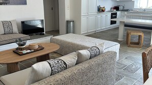 Ein modernes Wohnzimmer mit Sofa, Couchtisch und Fernseher. Es gibt eine Küchenzeile mit Schränken und Geräten.