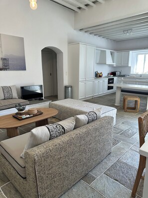 A modern living room with a sofa, coffee table, and TV. There's a kitchen area with cabinets and appliances. - Plakoto House (Exambela)