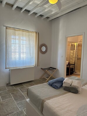 A bedroom with a bed, a radiator, a window with curtains, a towel rack, and a door leading to a bathroom. - Plakoto House (Exambela)