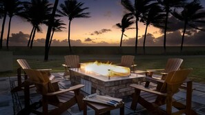 Un espacio al aire libre con fogata, sillas y vista a palmeras al atardecer.