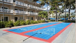 Una cancha de shuffleboard con mesa de billar, palmeras y un edificio de hotel al fondo.