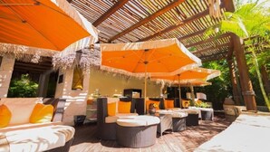 An outdoor lounge area with wicker furniture, orange umbrellas, and a thatched roof structure. - 1BR Suite – Beachfront Cancun, Pools & Ocean Views (Punta Sam)