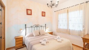 A bedroom with a bed, bedside tables, a chandelier, and a window with curtains. - El Almendro Cómpeta by Ruralidays (Málaga)