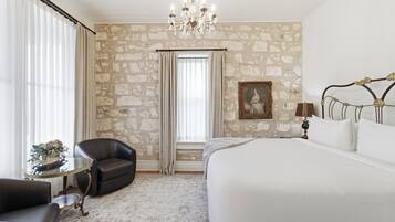 A bedroom with a large bed, a chandelier, a stone wall, a painting, and a small table with a lamp.