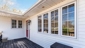 A covered porch with a black trash bin, a red door, and a window with a view of trees and a building.