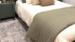 Un dormitorio ordenado con cama, dos mesitas de noche, una lámpara y una ventana con persianas.